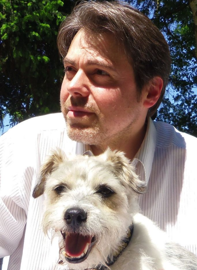 Mann mit kurzem Haar und Hemd, lächelt mit einem fröhlichen Hund neben sich.